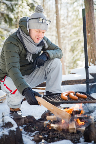 Man roasting sausages on campfire in forest by the lake, making a fire, grilling. Happy tourist exploring Finland. Beautiful sunny winter landscape, wood covered with snow.  © Suzi Media 
