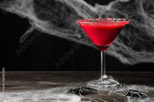 Close-up of glass with red halloween cocktail with spider, on wooden table, black background with spider web, horizontal, with copy space