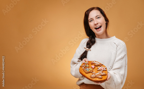 Happy pastry chef woman holding a wooden tray with gingerbread cookies for a traditional holiday christmas on a yellow background.