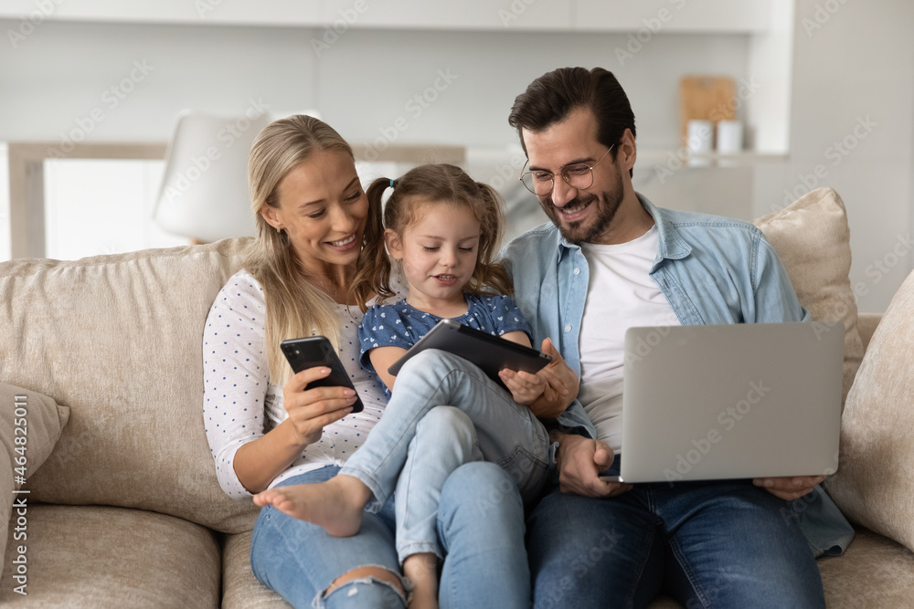 Happy bonding loving family couple parents with little preschool kid daughter holding different modern technology gadgets, spending time online sitting on comfortable couch addiction concept. Happy bonding loving family couple parents with little preschool kid daughter holding different modern technology gadgets, spending time online sitting on comfortable couch addiction concept.