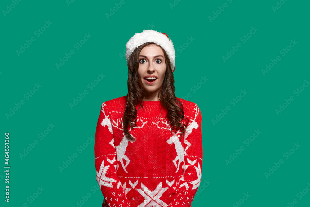Excited brunette has eyes and mouth wide open is amazed by gift or surprise, cant wait to unpack the box, wears red ugly sweater with deer and santa hat on green background. Christmas concept. Excited brunette has eyes and mouth wide open is amazed by gift or surprise, cant wait to unpack the box, wears red ugly sweater with deer and santa hat on green background. Christmas concept.