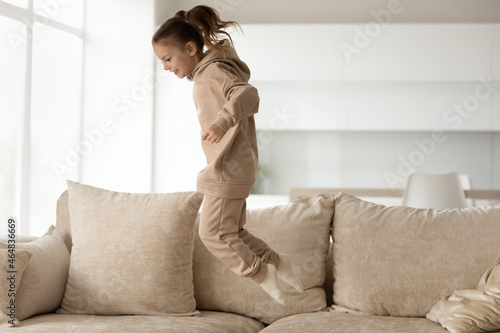 Full length joyful little 7s kid girl in comfortable loungewear jumping on sofa, having fun entertaining alone in living room. Happy small kid enjoying leisure active weekend pastime, playing at home. photo