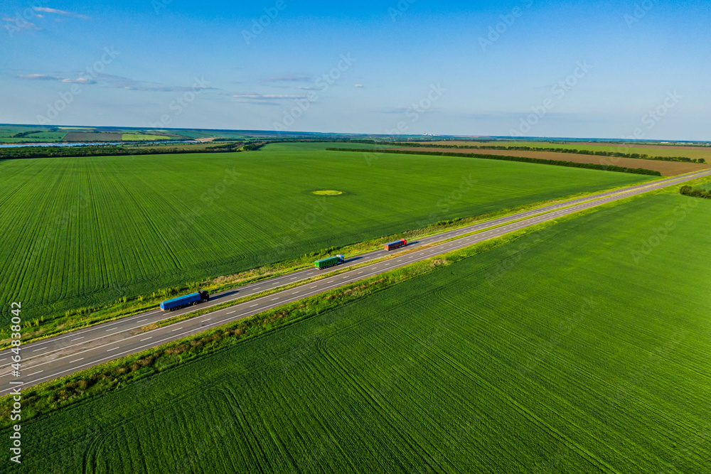 Obraz premium convoys with cargo. red green and blue trucks are driving on asphalt road along the green fields with goods. seen from the air. Aerial view landscape. drone photography.