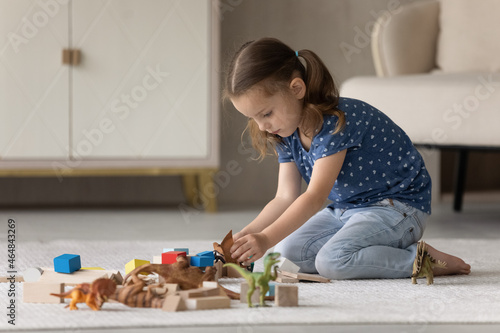 Happy adorable small preschool kid girl playing toys, sitting alone on floor carpet in modern living room. Joyful little 7s child having fun, involved in entertaining playtime activity at home.