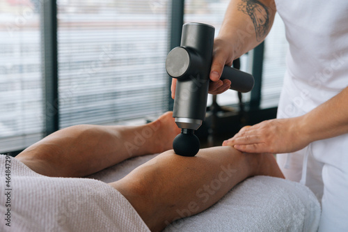 Close-up of unrecognizable professional male masseur massaging leg calf muscles using massage gun percussion tool of muscular athlete man, on spa treatment lying on back in massage table.