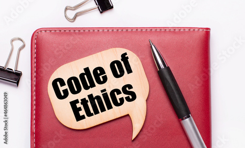 On a light background there are black paper clips, a pen, a burgundy notepad a wooden board with the text CODE OF ETHICS