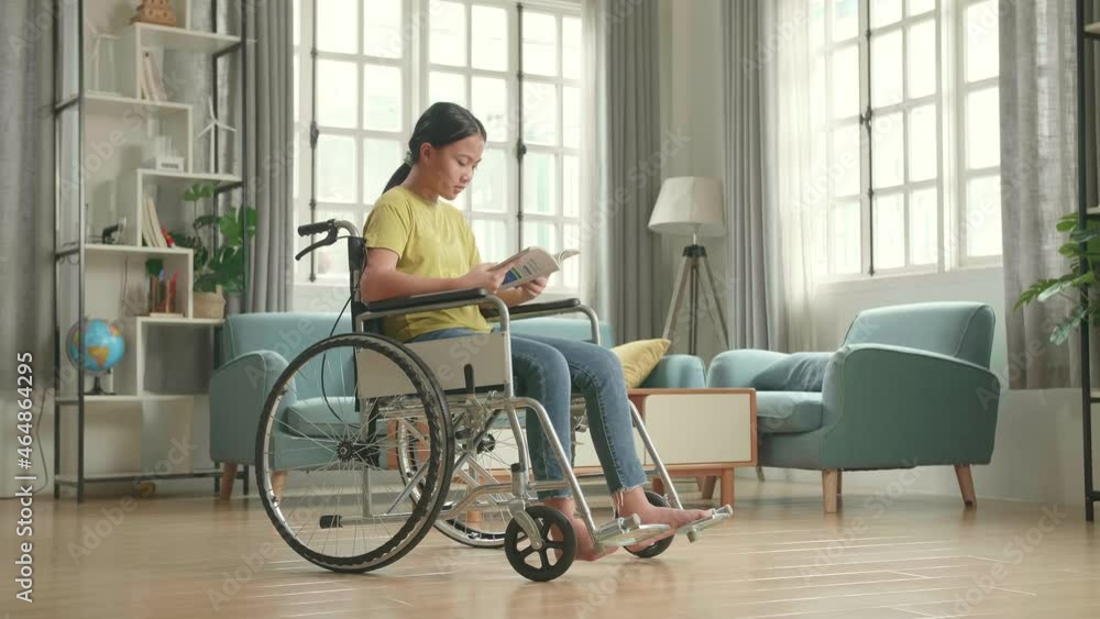 Asian Kid Girl Sitting In A Wheelchair And Reading Book At Home
