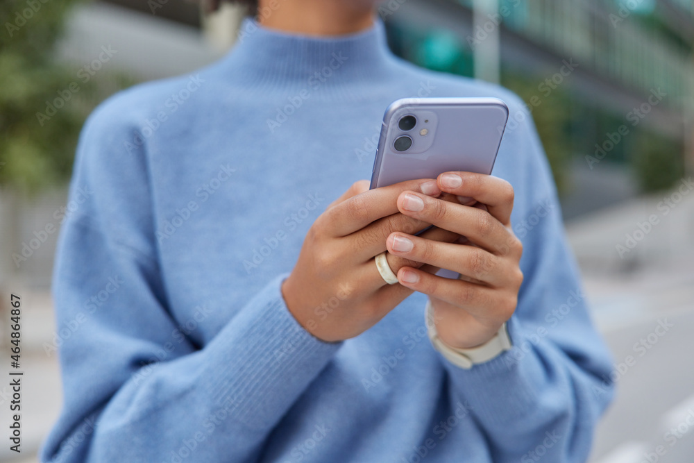 Cropped image of unrecognizable faceless woman in casual blue jumper holds mobile phone types sms message makes online shopping communicates in social networks poses outside uses free internet Cropped image of unrecognizable faceless woman in casual blue jumper holds mobile phone types sms message makes online shopping communicates in social networks poses outside uses free internet