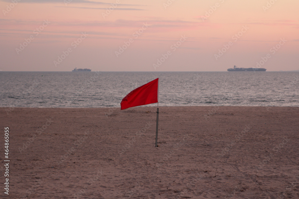 warning red flag on the beach of the Atlantic ocean with two ships on the horizon at sunset 