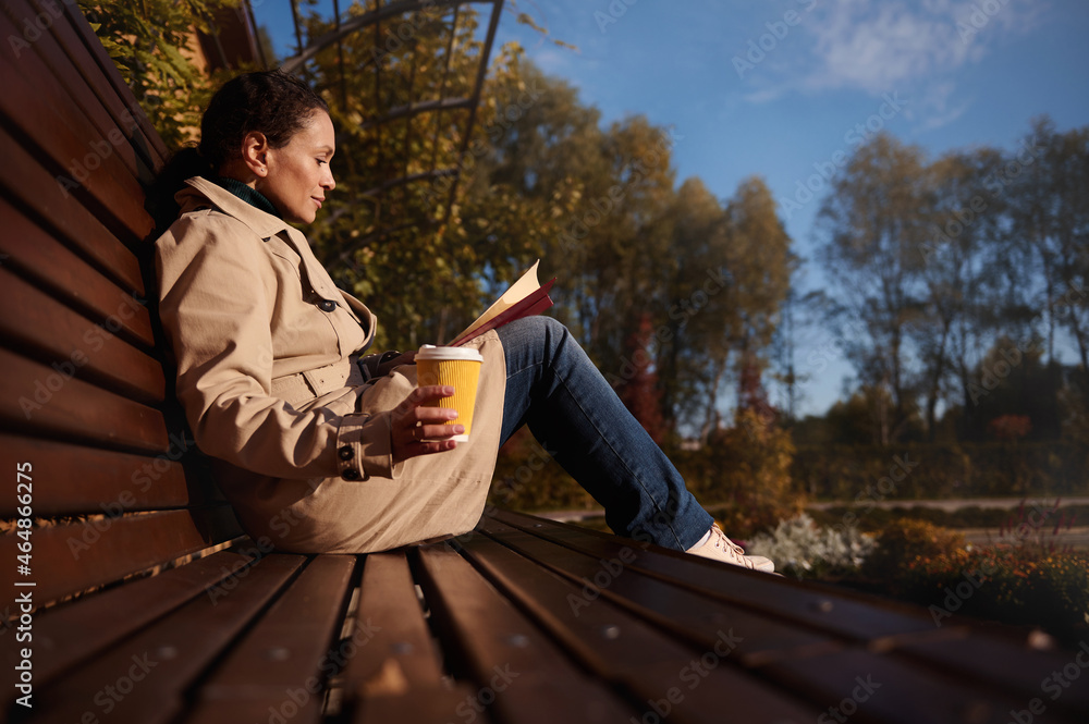 Fototapeta premium Relaxed pretty woman in casual denim and beige coat with a book and a takeaway paper cup of hot drink enjoying a warm sunny autumn weekend in country park, away from the hustle and bustle of the city