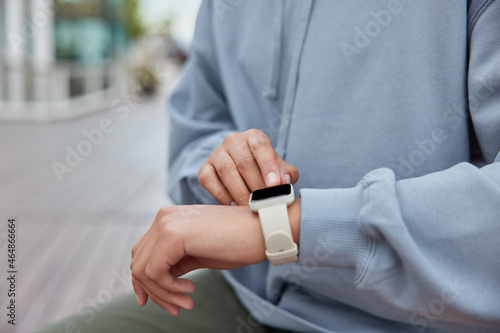 Cropped shot of unrecognizable sportswoman touches smartwatch screen monitors fitness results burned calories dressed in casual sweatshirt poses against blurred background checks notification