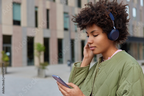 Profile shot of curly haired woman reads news via mobile phone wears wireless headphones listens audio track from playlist dressed in jacket stands against modern building checks social networks