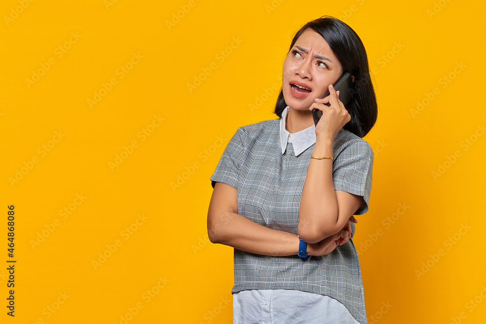 Portrait of angry young Asian woman while receiving incoming call on smartphone on yellow background Portrait of angry young Asian woman while receiving incoming call on smartphone on yellow background