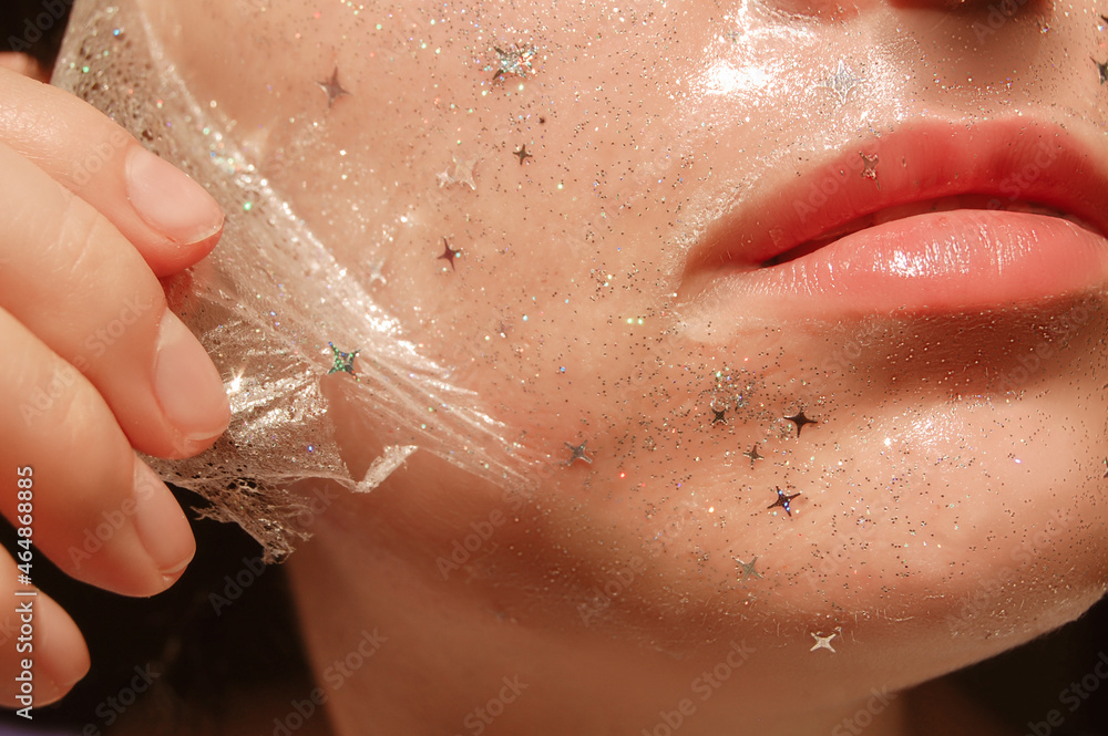 Fototapeta premium Young woman with healthy white pale skin removes purifying transparent peel off mask with shiny sparkling star glitter from her face. Trendy cosmetic beauty procedure.Skin care concept.