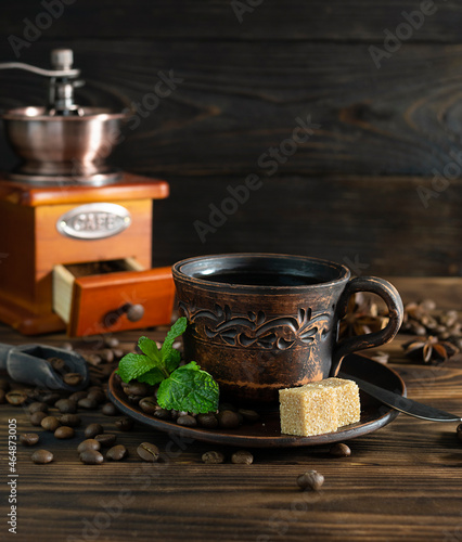 Black coffee in a clay cup on a dark wooden table. Coffee beans and mint leaves are on the saucer. Coffee grinder, star anise and cinnamon in the background