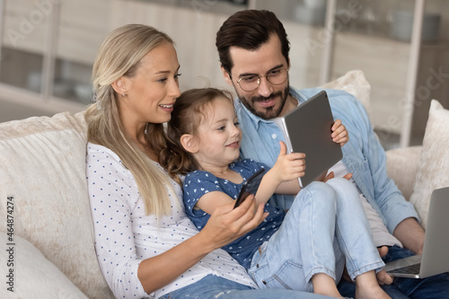 Addicted to modern technology happy young family couple with adorable little preschool child daughter using different gadgets, playing mobile games online or watching cartoons, spending time online.