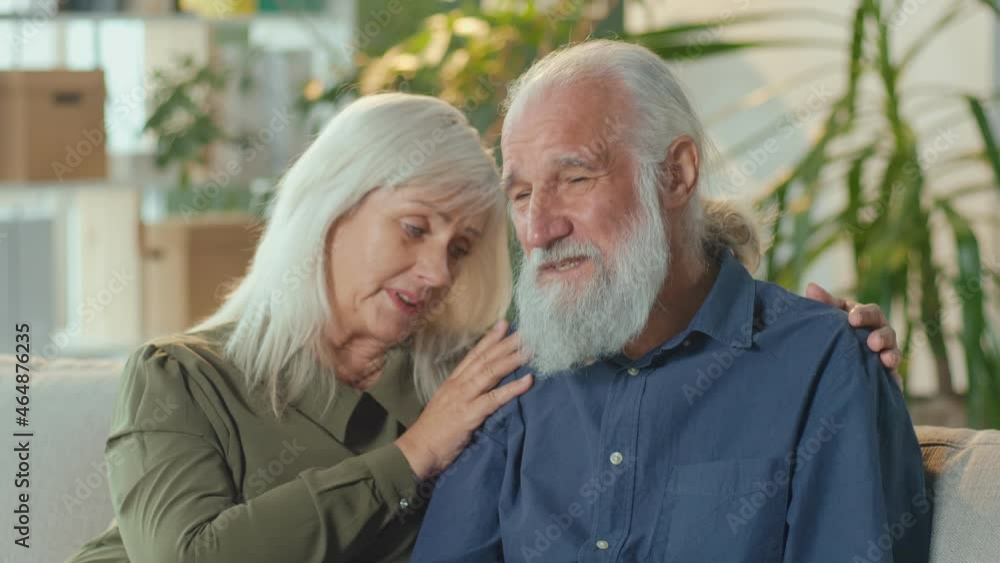 Depressed Elderly Couple Sitting on The Couch in the Living Room at Home. A Loving Wife Patiently Soothes, Consoles an Upset Husband. Family Relationships Concept. Mental Health. Depressed Elderly Couple Sitting on The Couch in the Living Room at Home. A Loving Wife Patiently Soothes, Consoles an Upset Husband. Family Relationships Concept. Mental Health.