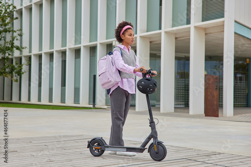 Female student rides to university by electric scooter carries rucksack poses against city buildings outdoors wears eleagant clothes. Millennial lifestyle urban transportation and modern technologies photo