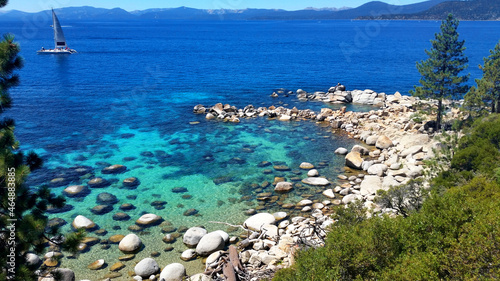 A wide shot of Lake Tahoe in the summer with a sailboat in the background on a sunny day and in the foreground a rocky blue cove, private beach