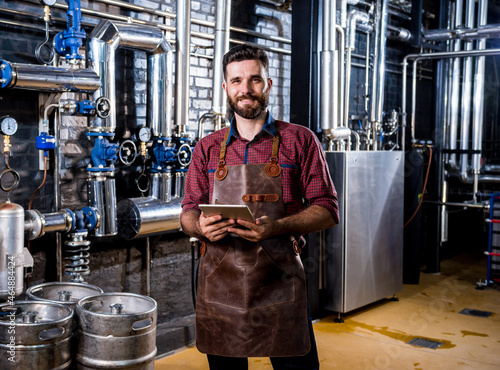 Young male brewer in leather apron supervising the process of beer fermentation at modern brewery factory