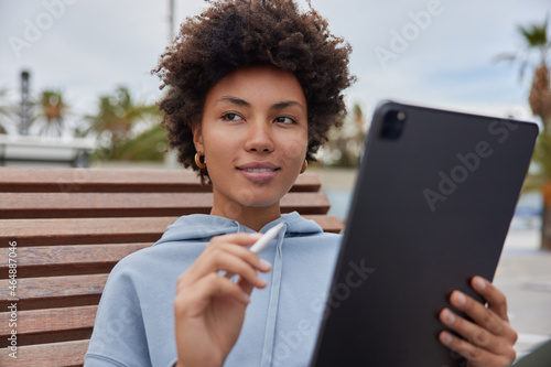 Dreamy satisfied woman with cutly hair holds modern tablet and stylus creats pictures or edits photos works outdoors wears casual hoodie poses at wooden bench duing daytime focused somewhere photo