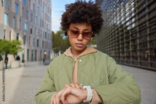 Serious stylish curly haired young woman looks at watch checks time waits for somone wears sunglasses casual hoodie and jacket poses at street among cityscrapers during daytime comes on meeting photo