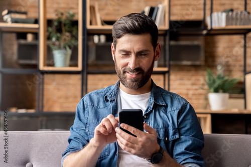 Smiling man using mobile phone scrolling network feed sitting on sofa at home 