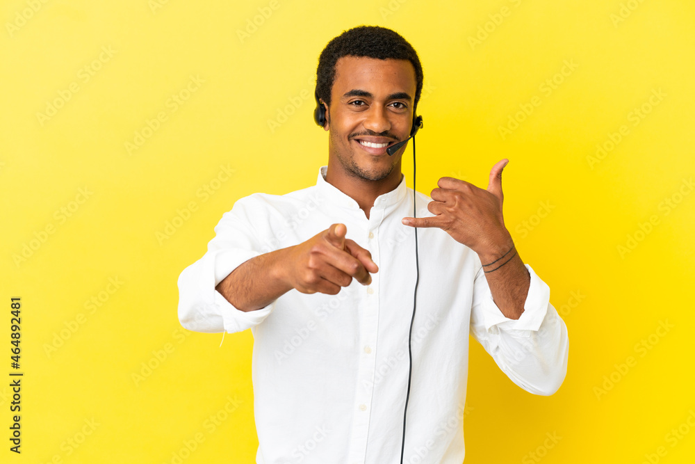 African American Telemarketer man working with a headset over isolated yellow background making phone gesture and pointing front African American Telemarketer man working with a headset over isolated yellow background making phone gesture and pointing front