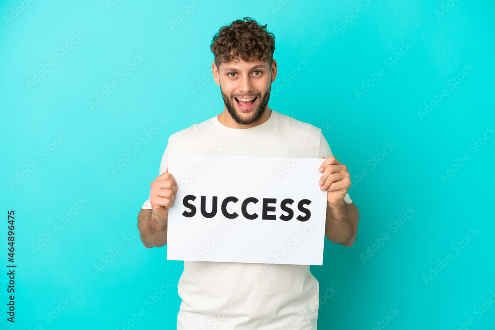 Young handsome caucasian man isolated on blue background holding a placard with text SUCCESS with surprised expression Young handsome caucasian man isolated on blue background holding a placard with text SUCCESS with surprised expression