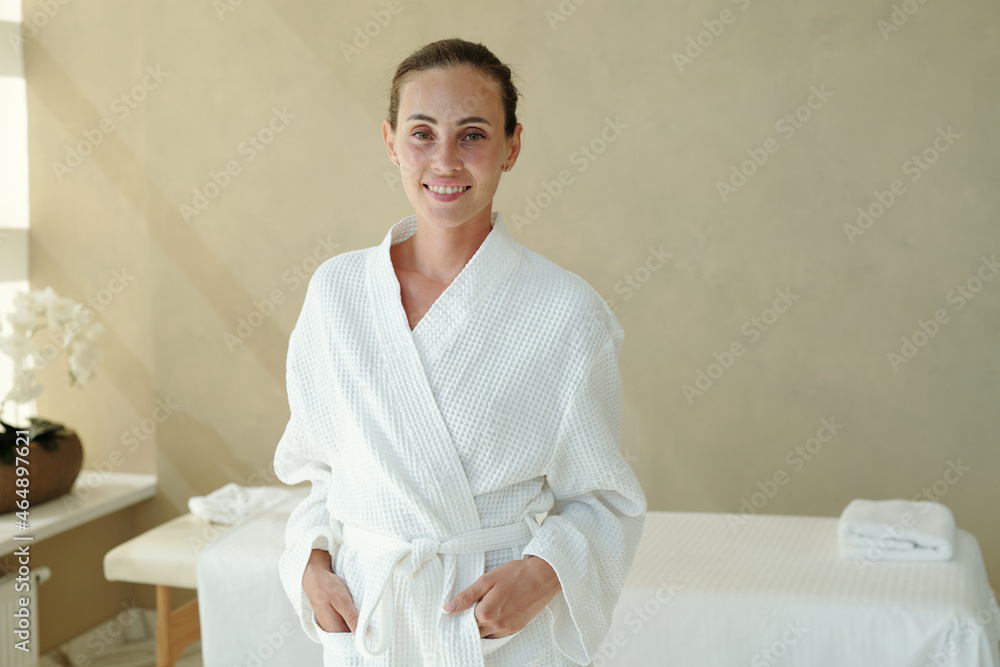 Young healthy woman in white bathrobe looking at you with smile while standing in spa salon Young healthy woman in white bathrobe looking at you with smile while standing in spa salon