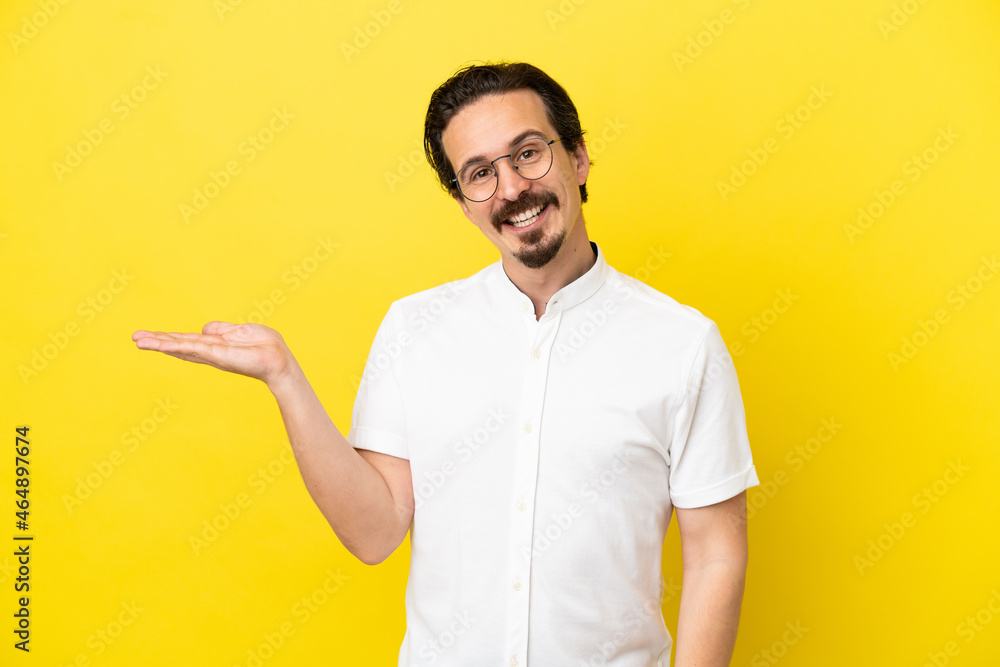Young caucasian man isolated on yellow background holding copyspace imaginary on the palm to insert an ad Young caucasian man isolated on yellow background holding copyspace imaginary on the palm to insert an ad