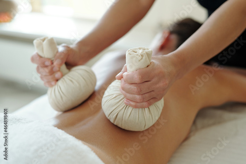 Masseuse pressing back of female client with two herbal compress balls during beauty procedure in spa salon