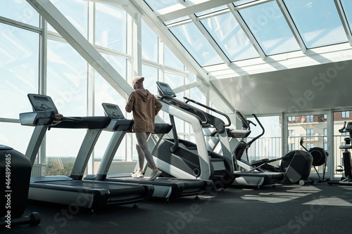 Back view of young active female running on treadmill in front of large window in contemporary leisure center