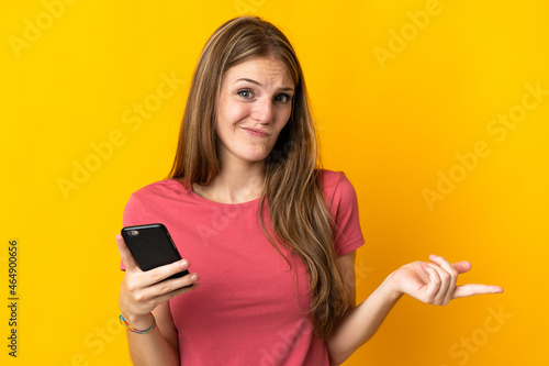 Young woman using mobile phone isolated on yellow background making doubts gesture while lifting the shoulders