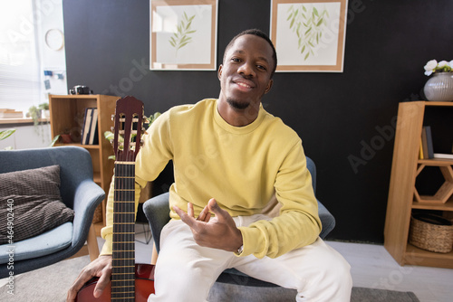 Happy young African teacher of music with guitar sitting in front of camera against home interior