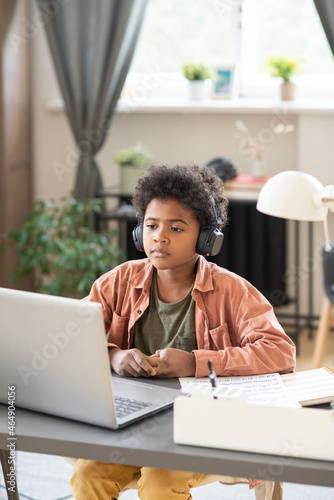 Serious schoolboy in headphones sitting in front of laptop and listening to teacher during online lesson