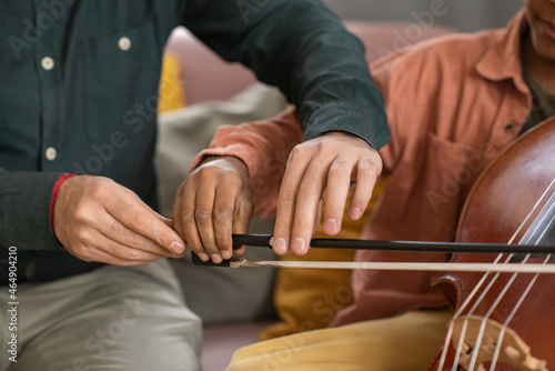 Hands of music teacher and his pupil playing cello while young man holding fiddlestick and helping little boy to glide it