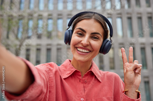 Positive carefree young European woman takes selfie makes peace gesture smiles happily wears headphones for listening music wears casual pink shirt poses against blurred building background. photo