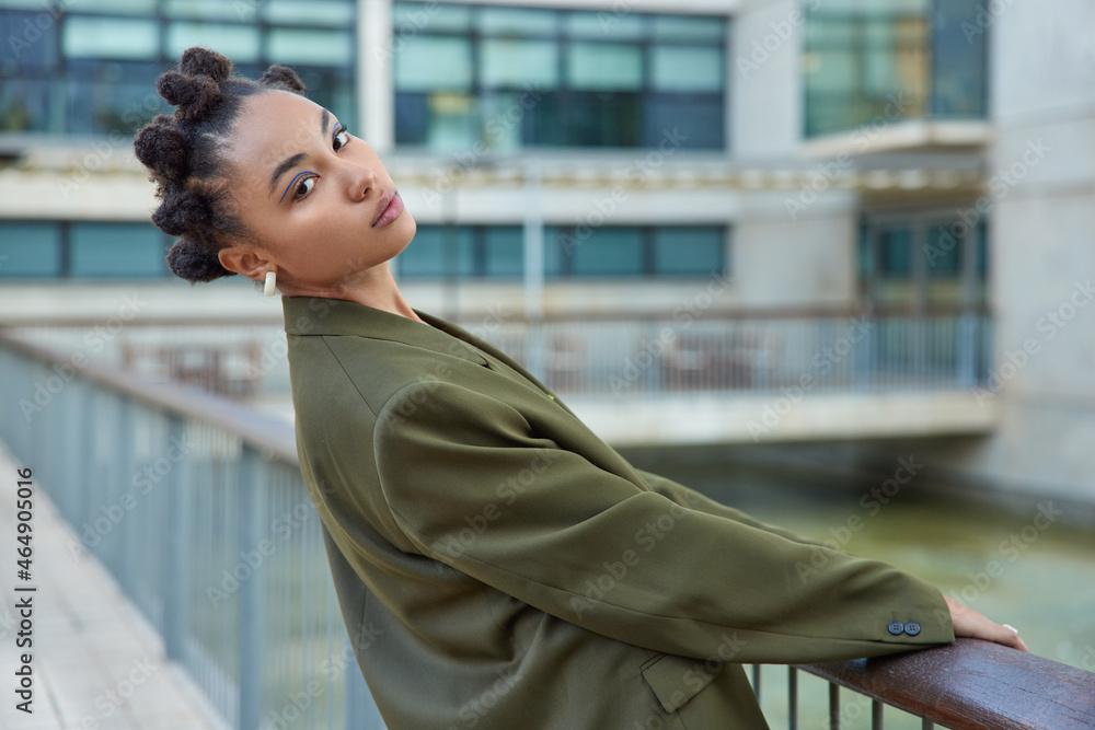 Sideways shot of fashionable young woman with bun haircut wears green jacket blue eyelines poses at bridge against blurred background has self confident expression. Stylish female poses for photo Sideways shot of fashionable young woman with bun haircut wears green jacket blue eyelines poses at bridge against blurred background has self confident expression. Stylish female poses for photo