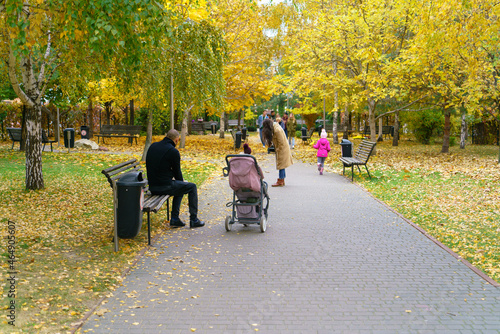 Young father sits on a park bench next to a baby carriage in a city park on an autumn day