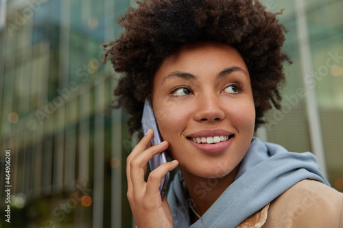 Close up shot of happy curly haired woman smiles gently concentrated away makes phone call talks with friend wears hoodie keeps smartphone near ear. Beautiful female has friendly conversation