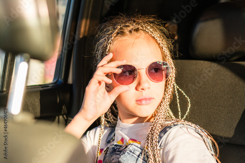 Serious teenager girl with afro-braids holds sunglasses with her hand and looks out the window while sitting in the car. Road trip