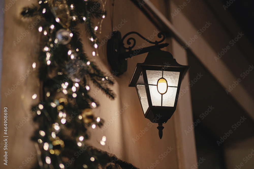 Stylish christmas lantern and spruce branches lights illumination on old building in evening. Atmospheric magic time. Christmas festive decor for winter holidays. Merry Christmas!