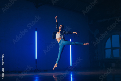 Young woman in casual style - jeans and leather jacket doing ballet in modern studio. Attractive ballerina practices in choreography alone