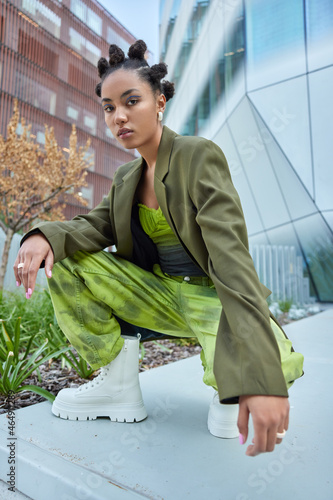 Outdoor shot of stylish woman with hairstyle vivid makeup green fashionable clothes and white boots poses against modern buildings in city. Trendy hipster girl spends free time in urban setting photo