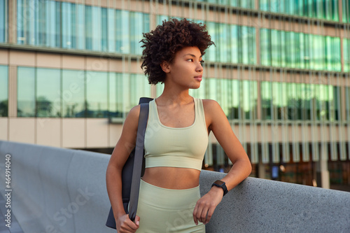 Horizontal shot of sporty thoughtful woman with curly hair dressed in tracksuit wears smartwatch on wrist carries karemat rests after successful fitness training poses against urban setting.