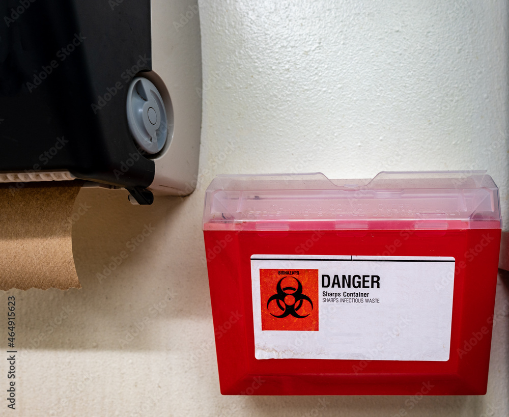 Red biohazard sharps waste container mounted to the wall of a public restroom - obrazy, fototapety, plakaty 