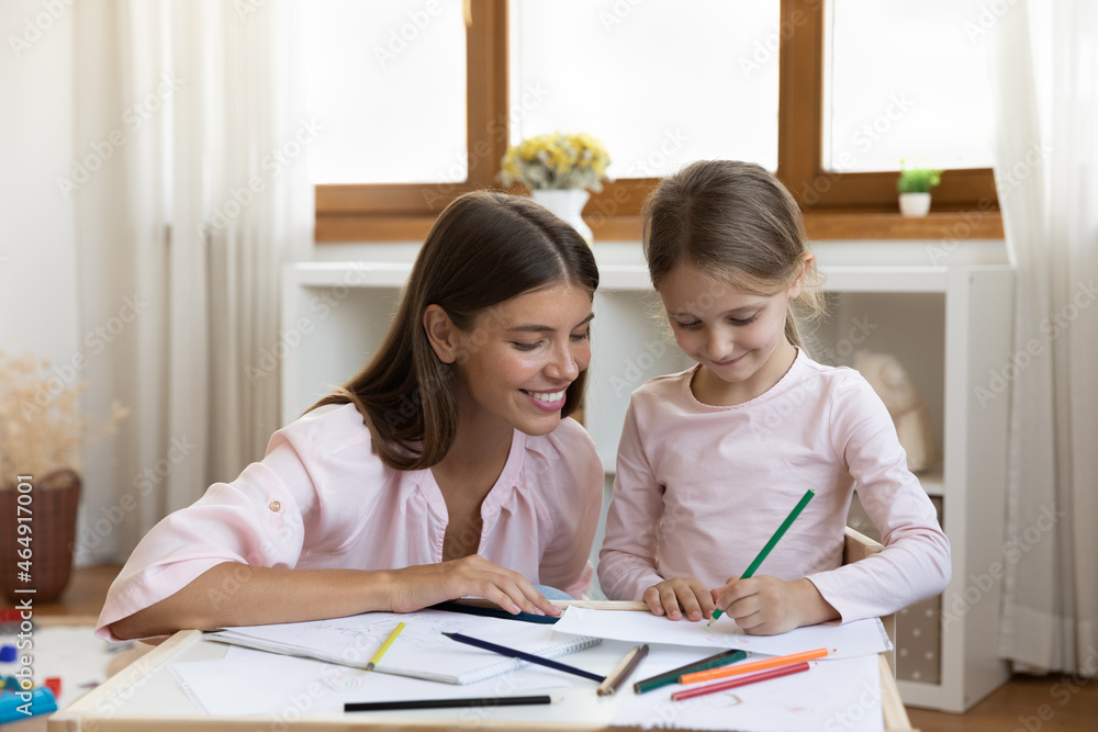 Smiling attractive millennial mother or babysitter enjoying hobby activity with small child girl, drawing or coloring pictures in paper album, single parenting, creative skills development concept. Smiling attractive millennial mother or babysitter enjoying hobby activity with small child girl, drawing or coloring pictures in paper album, single parenting, creative skills development concept.