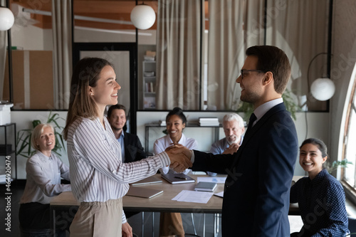 Acknowledgement and recognition. Grateful millennial male boss team leader shake hand of happy young female intern hire on job. Smiling diverse staff watching smiling director welcoming new colleague