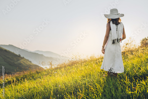 a woman wearing a hat and white dress on a hillside and looking at the horizon at sunset. concept of nature and environment.